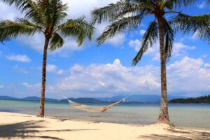 Borneo Gaya island-2 | Kenzan Travel Hammock stretched between two palm trees on a sandy beach with calm turquoise water and blue sky behind.
