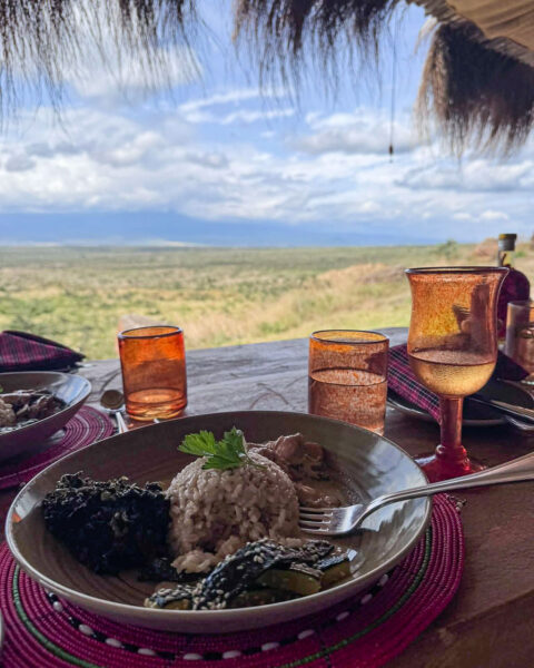 Tanzania safari Ellas familj-15 | Kenzan Travel Outdoor dining scene: plated meal (rice with greens and sesame veggies) on a wooden table with amber glasses, desert landscape in the distance under a thatched roof.