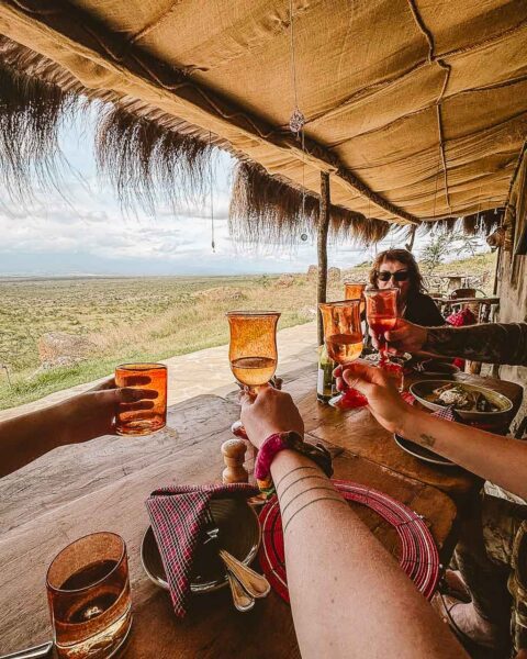 Tanzania safari Ellas familj-2 | Kenzan Travel People toasting with orange wine glasses at a rustic outdoor table, overlooking a dry landscape.