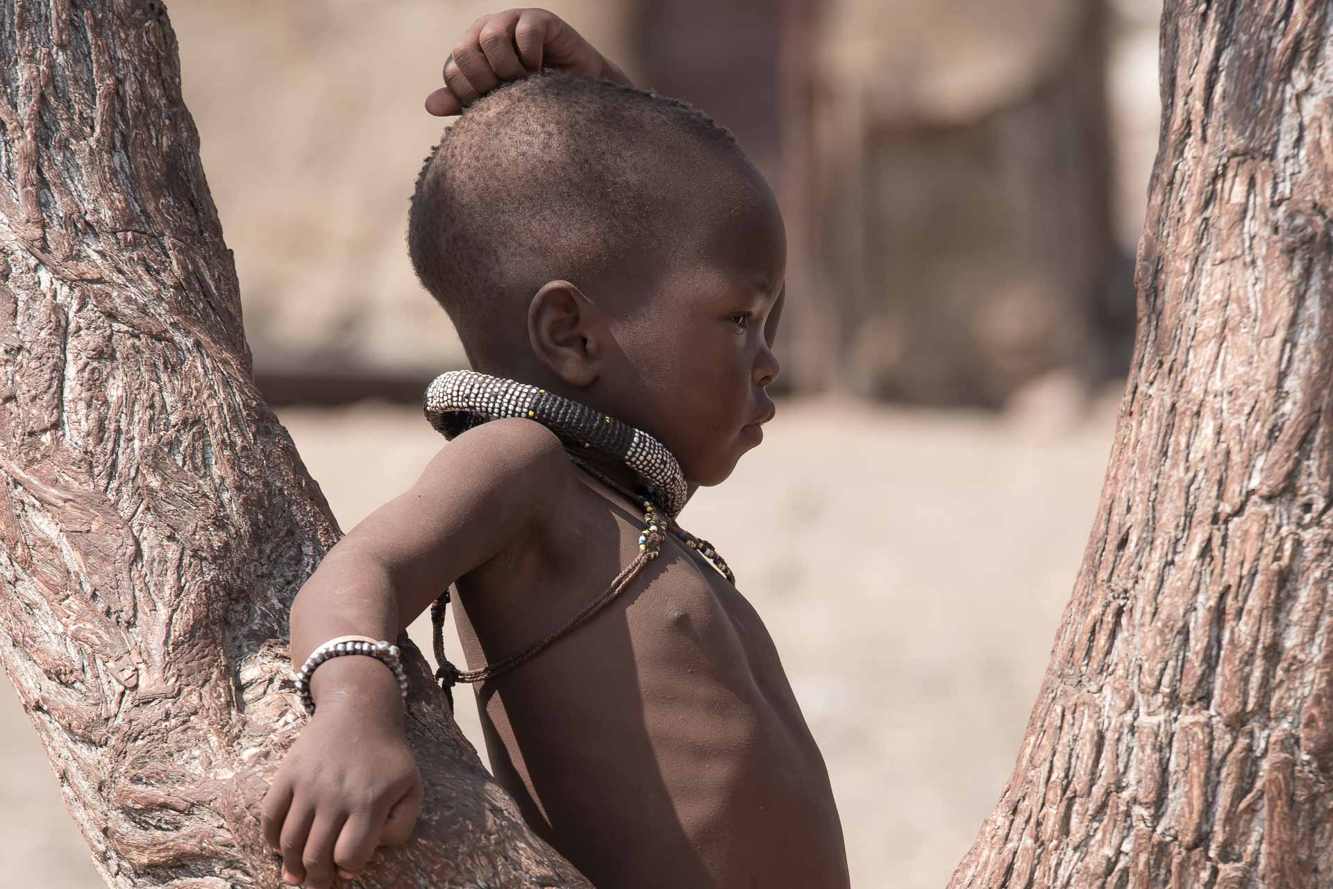 Namibia Himba | Kenzan Travel Young child with beaded necklaces leaning between two tree trunks in a dusty outdoor setting.