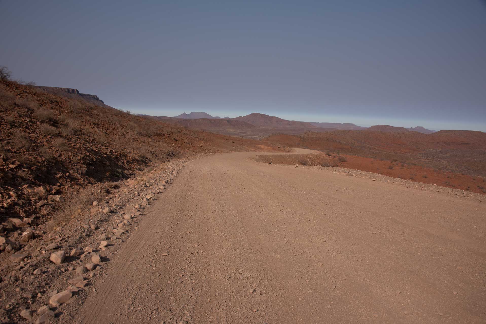 Namibia Sceleton Coast | Kenzan Travel Dusty unpaved road winding through a barren desert with rocky hills on the left and distant red mesas on the horizon under a clear blue sky.
