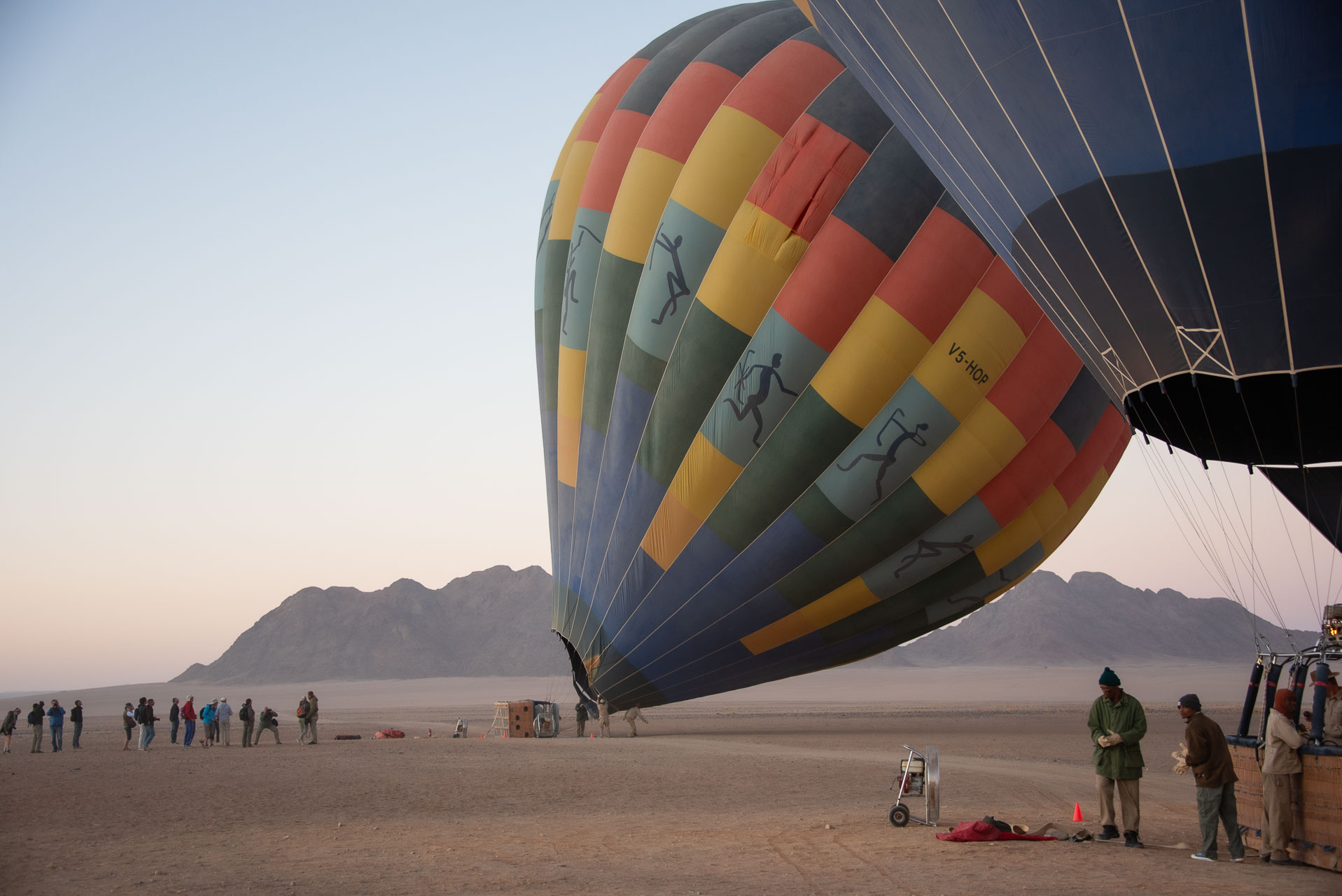 Namibia Sossusvlei ballongfärd | Kenzan Travel People stand in a line on a sandy beach as a large, colorful hot air balloon is prepared for inflation at sunset. Mountain range in the background.