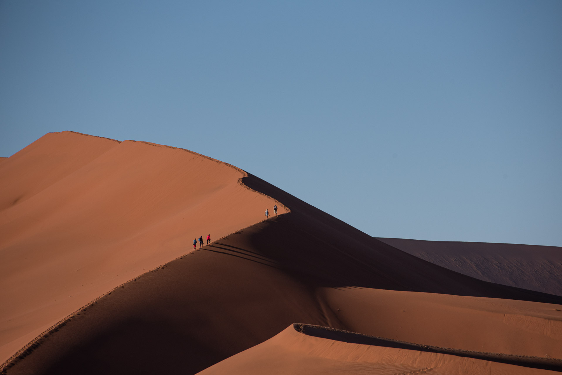 Namibia Sossusvlei sanddynor-2 | Kenzan Travel Hikers walking along the ridge of a large orange sand dune against a clear blue sky.